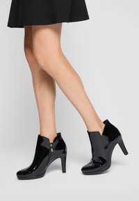Bottines en cuir verni noir avec un talon haut et un bout arrondi. Comprend une fermeture éclair sur le côté et une découpe en forme de V au niveau de la cheville.
