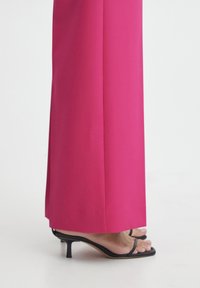 Fuchsia weit geschnittene Hose aus glattem Stoff, kombiniert mit schwarzen Riemchensandalen mit hohen Absätzen, die eine elegante Silhouette zeigen.