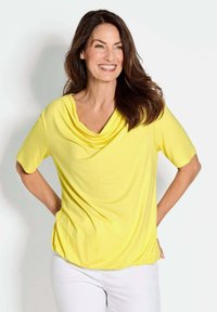 Gelbes, drapiertes Oberteil mit kurzen Ärmeln und einem Cowl-Ausschnitt, aus einem weichen Stoff gefertigt. Kombiniert mit weißen Hosen, vor einem hellen Hintergrund.