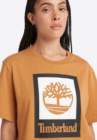 Camiseta de algodón de color mostaza con mangas cortas que presenta un gráfico cuadrado negro con un logotipo de un árbol y el texto "Timberland" debajo.