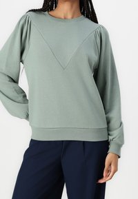 Sudadera verde claro con cuello redondo, con hombros abullonados y detalles de costura en V. Los puños y el dobladillo acanalados añaden textura.