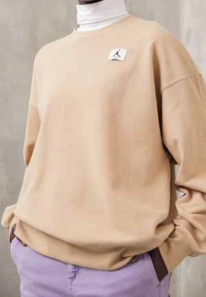 Personne portant un sweat-shirt beige sur un col roulé blanc avec un petit patch logo Jumpman noir, associé à un pantalon violet clair, mains dans les poches.