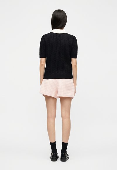Femme portant un haut texturé à manches courtes noir avec col blanc, un short texturé rose clair, des chaussettes noires et des chaussures noires, debout de dos.