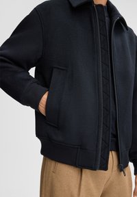 Black fleece jacket with a stand collar, side pockets, ribbed cuffs, and a textured zipper placket. Worn over a dark shirt and tan trousers.