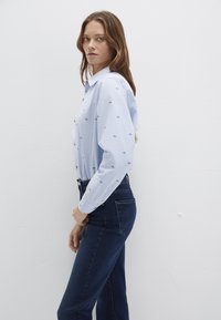 Light blue button-up shirt with small floral embroidery, long sleeves, and a collar, paired with dark blue high-waisted jeans.