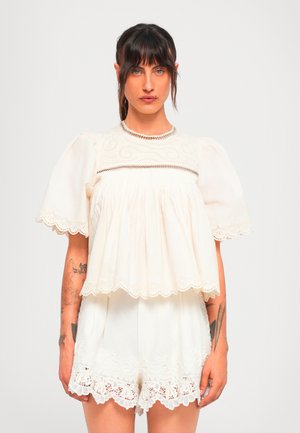 Woman with dark hair and tattoos wears white embroidered short-sleeve blouse and matching lace-trimmed shorts, standing against plain background.