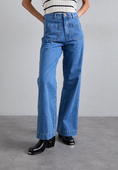 High-waisted blue denim jeans with wide legs, featuring two back pockets and stitched hem. Paired with black heeled clogs.