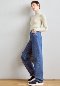 Beige ribbad polotröja, höga blå denimjeans och svarta sportskor med vita detaljer. Modellen poserar mot en neutral bakgrund.