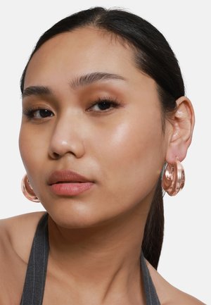 Boucles d'oreilles créoles en or rose avec une finition lisse et brillante, présentant un design arrondi et sculptural. Portées sur l'oreille d'un mannequin.