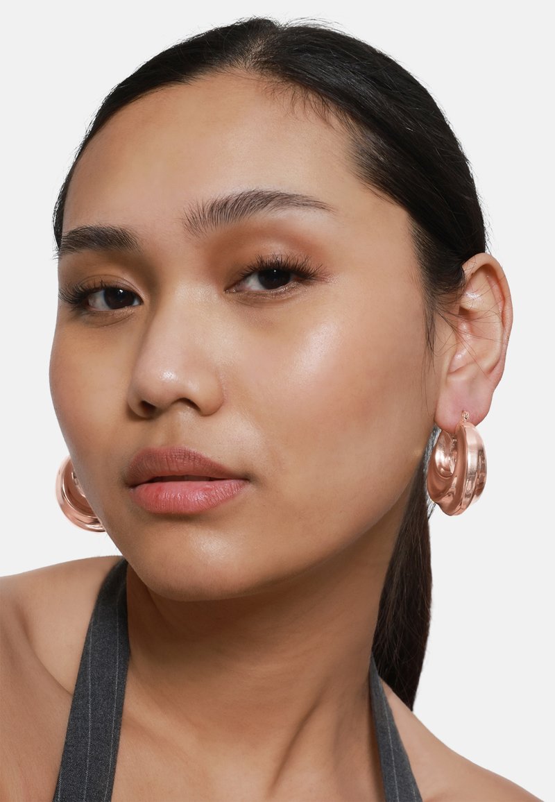 Boucles d'oreilles créoles en or rose avec une finition lisse et brillante, présentant un design arrondi et sculptural. Portées sur l'oreille d'un mannequin.