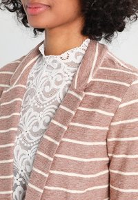 Randig, lätt, vävd cardigan i dammros med vita ränder, med en notch-krage och en texturerad yta, lagrad över en spetsblus.