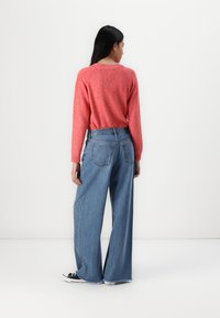 Pull en maille texturée rouge associé à un jean bleu en denim à jambes larges avec des ourlets effilochés et des poches arrières, porté avec des baskets noires.
