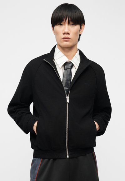 Νεαρός άνδρας με ίσια μαύρα μαλλιά, φορώντας μαύρο μπουφάν με φερμουάρ πάνω από άσπρο πουκάμισο και μαύρη γραβάτα, με τα χέρια στις τσέπες του μπουφάν.