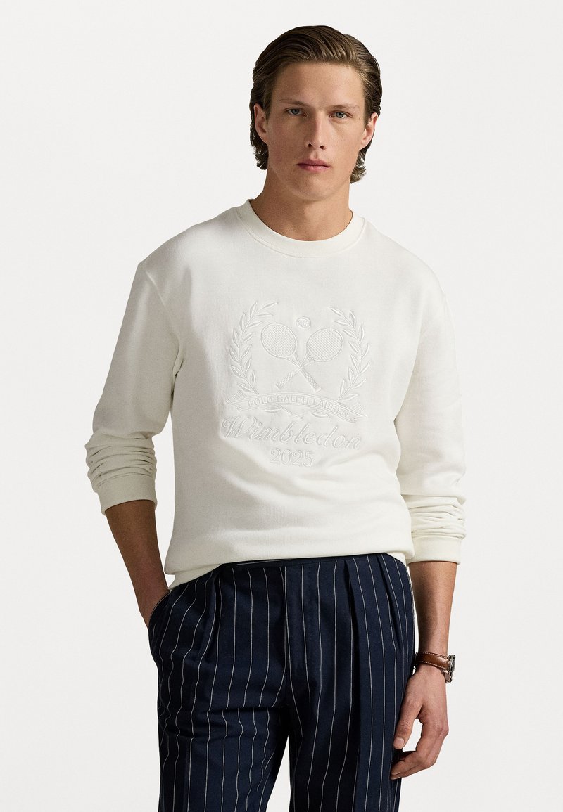 Hanorac alb cu un motiv brodat de tenis și textul "Wimbledon 2025", asortat cu pantaloni navy cu dungi subțiri. Textură moale, design cu guler rotund.