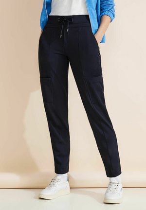 Navy trousers with elastic waistband, drawstring, and multiple pockets; made of smooth material. Paired with white sneakers.