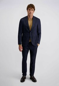 Mann steht in einem marineblauen Blazer und Hose mit einem senfgelben Pullover und dunkelbraunen Lederloafers vor einem schlichten hellen Hintergrund.