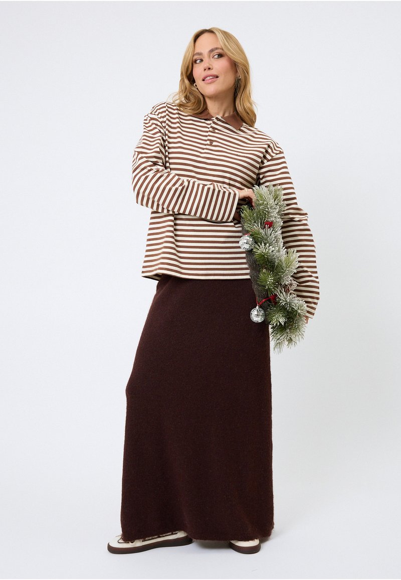 Svītrains brūnas un krēmkrāsas garām piedurknēm tops ar apkakli, kopā ar garu tumši brūnu svārku, turot dekorētu vītni.