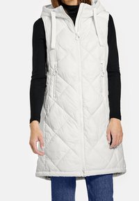 Pikowana biała bezrękawnik puchowy z kapturem, dwoma zapinanymi na zamek kieszeniami z przodu oraz pełnym zapięciem na zamek. Nosi się go na czarną bluzkę z długim rękawem.