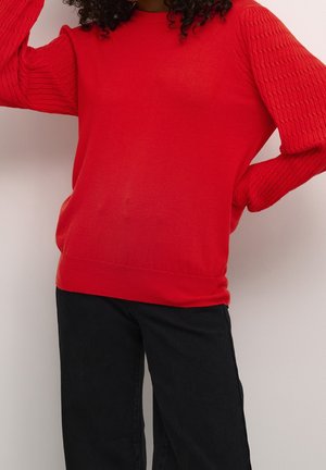 Personne portant un pull rouge vif avec des manches texturées et un pantalon noir, debout avec une main sur la hanche contre un mur blanc.
