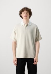 Πόλο μπλουζάκι σε ανοιχτό κρέμα με λευκό γιακά, κοντά μανίκια και άνετη εφαρμογή. Διαθέτει κουμπιά μπροστά και διακριτή υφή.