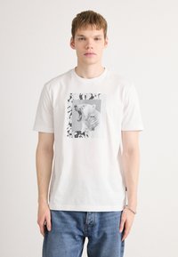 Fehér pamut póló, amelyen egy üvöltő jegesmedve grafika látható szürkeárnyalatban, absztrakt fekete-fehér mintákkal keretezve. Lazán illeszkedő fazon.