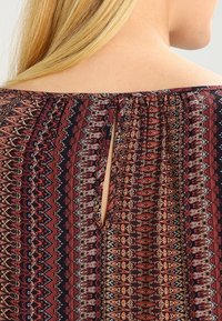 Tessuto a motivi con ricchi colori rosso, blu navy e crema; presenta un'apertura nella parte posteriore del collo; materiale liscio e leggero.
