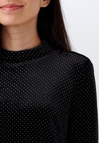 Černý, strukturovaný top s vysokým límcem a jemnými ozdobami. Má měkkou tkaninu s malými lesklými tečkami po celé ploše.