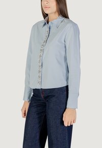 Camicetta azzurro chiaro a maniche lunghe con colletto, caratterizzata da bottoni decorativi a forma di fiore in argento lungo la parte frontale. Vestibilità classica con texture liscia.
