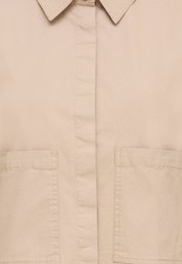 Smėlio spalvos marškiniai su sagomis, turi apykaklę, du priekiniai kišeneliai ir lygios audinio tekstūros. Paprastas dizainas su tiesiomis siūlėmis.