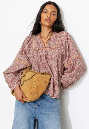 Une femme portant un chemisier ample à motifs et un jean bleu tient un sac à main en daim beige sur fond blanc.