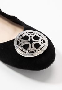 Sorte ruskinds flade sko med et stort, sølvfarvet dekorativt emblem med udskårne mønstre på tæerne. Glat indvendigt foer.