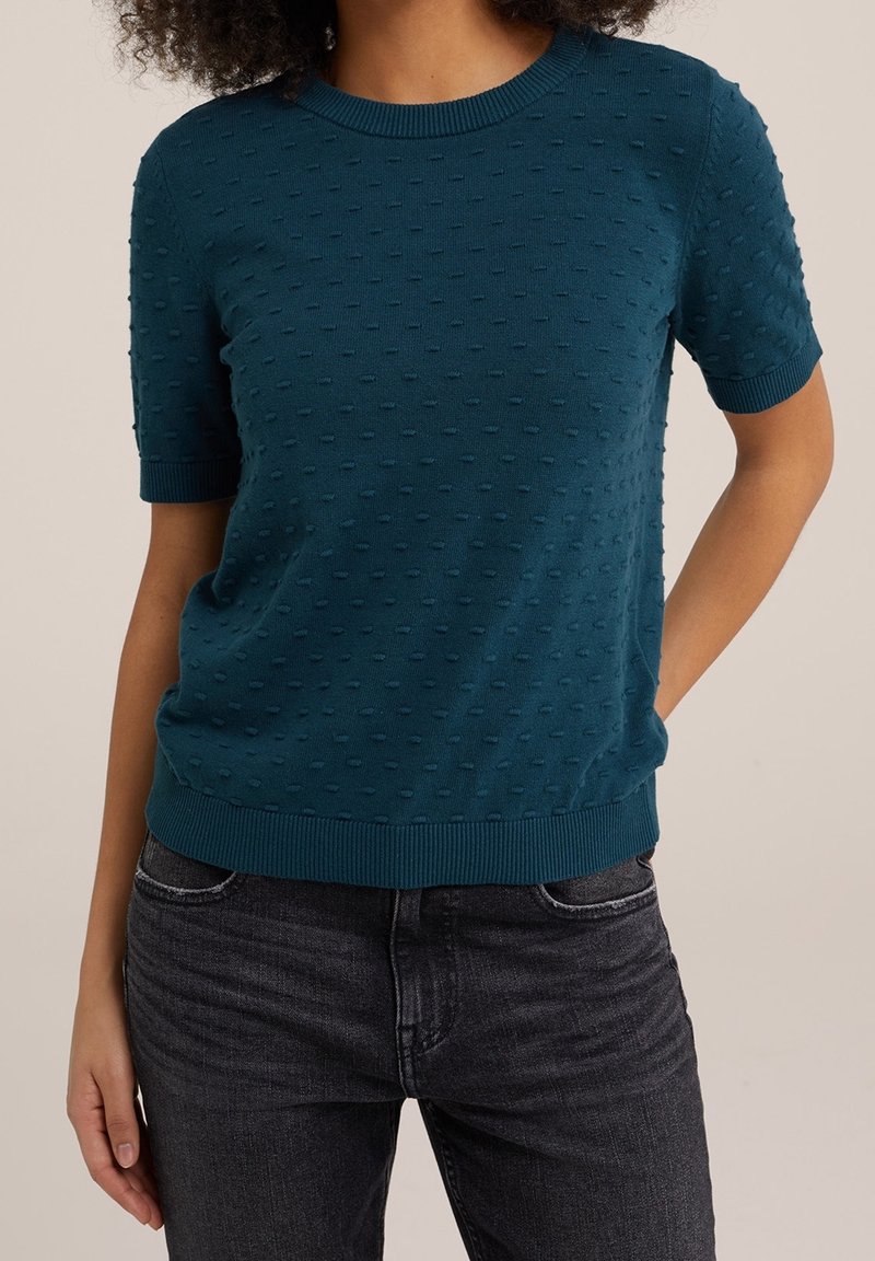 Haut en tricot texturé à manches courtes de couleur sarcelle avec de petits points en relief, col rond côtelé et poignets, porté avec un jean noir.
