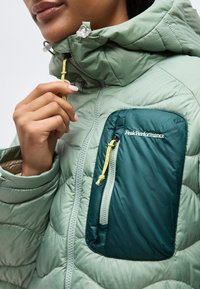 Personne ajustant la fermeture éclair d'une veste matelassée vert clair avec une poche poitrine vert foncé portant l'étiquette "Peak Performance".