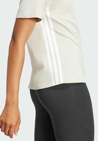 Helles beige T-Shirt mit kurzen Ärmeln, das drei weiße vertikale Streifen an der Seite aufweist, kombiniert mit schwarzen Leggings. Weicher Stoff.