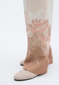 Persiku krāsas apavi ar gludu tekstūru, kuriem ir baltas ziedu raksti un kontrastējoša brūna papēža daļa, atklājot elegantu siluetu.