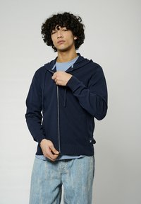 Sweat à capuche zippé bleu marine en tissu texturé avec un cordon de serrage. Associé à une chemise bleu clair, porté avec un jean en denim clair déchiré.