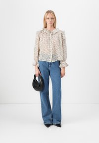 Blouse florale transparente avec un col à volants, fermeture à boutons sur le devant et manches longues ; assortie à un jean flare bleu clair et un sac à main noir.