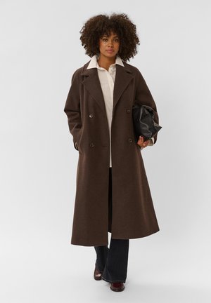 Femme aux cheveux bouclés portant un long manteau marron croisé, une chemise blanche, un pantalon noir, des chaussures bordeaux et tenant un sac noir.