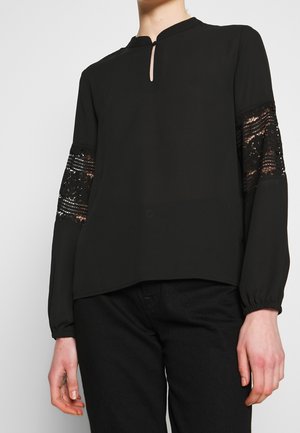 Woman wearing a black long-sleeve blouse with lace panels on the sleeves and a small keyhole neckline.