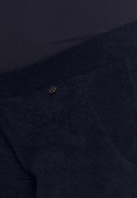 Calças de tecido azul marinho, com uma textura suave e um corte relaxado, apresentando um bolso de remendo e um pequeno botão metálico redondo.