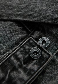 Tessuto denim nero con superficie testurizzata, con due bottoni a pressione neri etichettati "DENIM" e "OMBRE", posizionati in alto a sinistra.