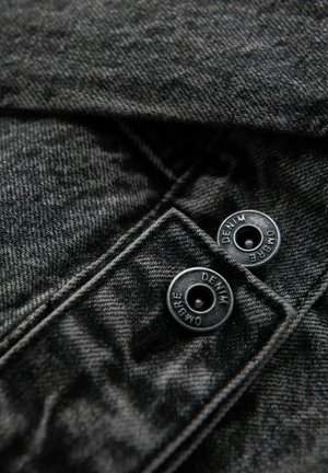 Schwarzer Denim-Stoff mit strukturierter Oberfläche, ausgestattet mit zwei schwarzen Druckknöpfen, die mit "DENIM" und "OMBRE" beschriftet sind, und sich oben links befinden.