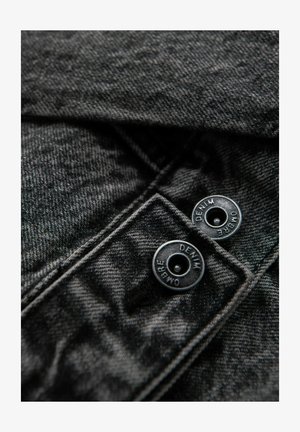 Schwarzer Denim-Stoff mit strukturierter Oberfläche, ausgestattet mit zwei schwarzen Druckknöpfen, die mit "DENIM" und "OMBRE" beschriftet sind, und sich oben links befinden.