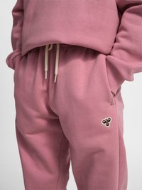 Nahaufnahme einer Person, die pinke Jogginghosen und ein Sweatshirt trägt, mit Händen in den Taschen, mit einem Bienenlogo am linken Oberschenkel.