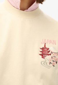 Felpa crema, colletto a coste, con ricamo di una pagoda giapponese, fiori di ciliegio e la scritta "GIAPPONE" in rosa. Texture morbida, design casual.