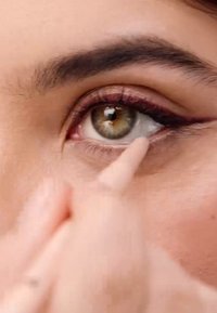 Eyeliner a matita applicato sulla palpebra inferiore, che mette in evidenza le tonalità verdi e marroni degli occhi. La pelle ha una texture liscia, con accenti di trucco sottili.
