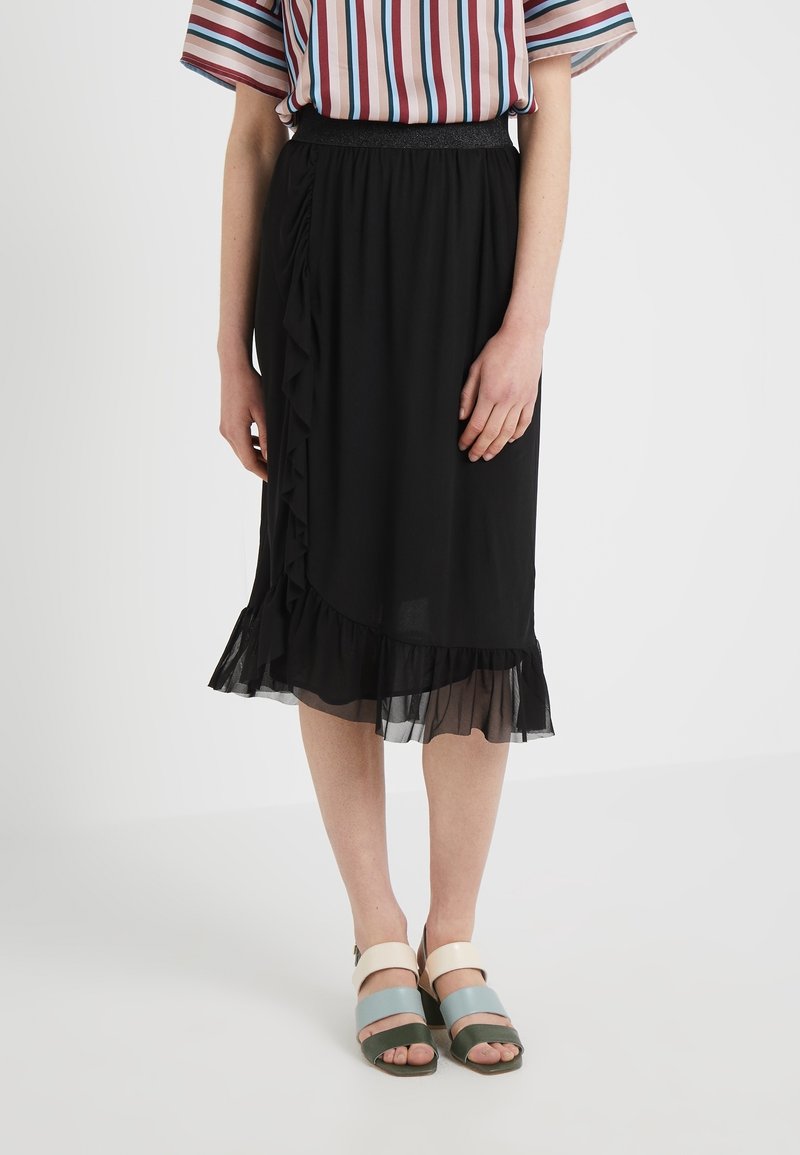 Schwarzer Midi-Rock mit Rüschendetails und transparentem Saum. Kombiniert mit mehrfarbigen Riemchensandalen. Modell trägt eine gestreifte Bluse.