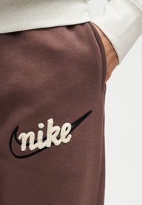 Χέρι στην τσέπη φορώντας καφέ φόρμα Nike με λευκό λογότυπο Nike και μαύρο σβούρο, συνδυασμένη με ανοιχτό κρεμ μακρυμάνικο τοπ.