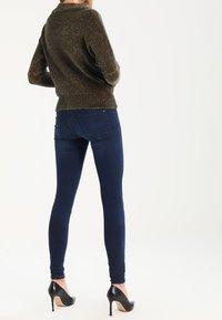 Sweater texturé vert foncé avec un ourlet côtelé, associé à un jean skinny bleu foncé et des chaussures à talons hauts noires, vu de dos.