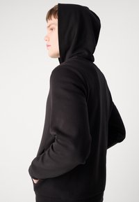 Svart hettegenser laget av mykt stoff. Har en enkel design, lange ermer og lommer foran. Glatt tekstur med en avslappet passform.